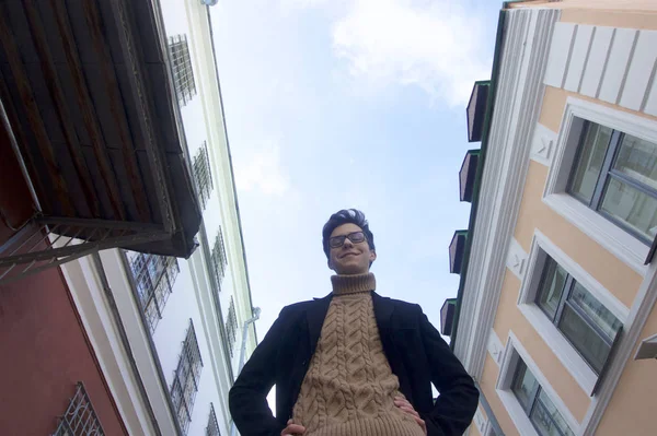 Un jeune homme souriant pose debout dans une rue piétonne de la vieille ville. Habillé d'un manteau léger noir, des lunettes à bord noir. En contre-jour, l'angle par en bas . — Photo