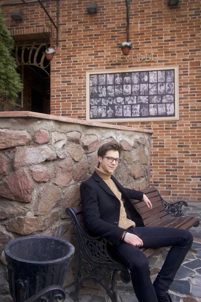 Un jeune homme au visage pensif est assis sur un banc. Habillé d'un manteau léger noir, lunettes à bord noir . — Photo