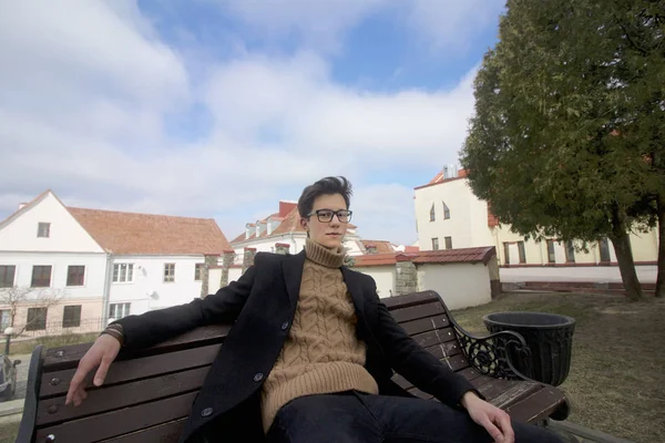 Un jeune homme au visage pensif est assis sur un banc. Habillé d'un manteau léger noir, lunettes à bord noir . — Photo