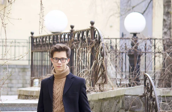 Un jeune homme au sourire pose, parmi les éléments de l'architecture de la vieille ville. Habillé d'un manteau léger noir, lunettes à bord noir . — Photo
