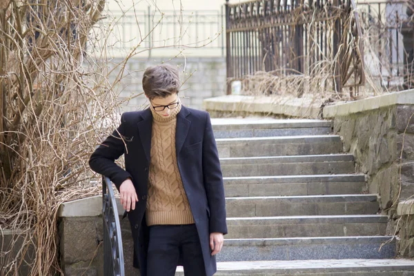 Un jeune homme au visage cher posant parmi les éléments de l'architecture de la vieille ville. Habillé d'un manteau léger noir, lunettes à bord noir . — Photo