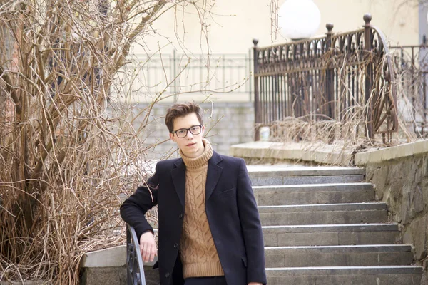 Un jeune homme au visage cher posant parmi les éléments de l'architecture de la vieille ville. Habillé d'un manteau léger noir, lunettes à bord noir . — Photo