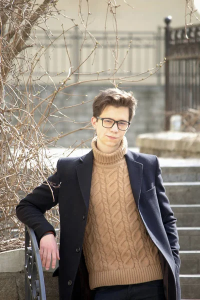 Un jeune homme au visage cher posant parmi les éléments de l'architecture de la vieille ville. Habillé d'un manteau léger noir, lunettes à bord noir . — Photo