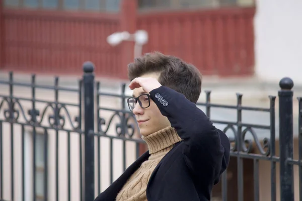 Un jeune homme au visage pensif est assis sur un banc. Habillé d'un manteau léger noir, lunettes à bord noir . — Photo