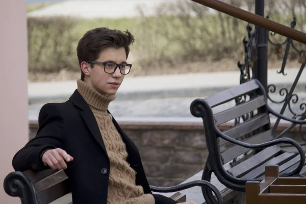 Un jeune homme au visage pensif est assis sur un banc. Habillé d'un manteau léger noir, lunettes à bord noir . — Photo