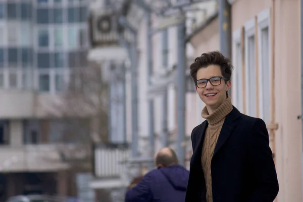 Un jeune homme posant avec un sourire, au milieu du paysage urbain. Habillé d'un manteau léger noir, lunettes à bord noir . — Photo