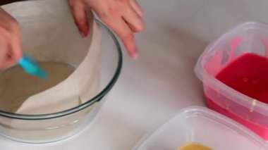 Bir kadın bir cam pişirme çanağı gıda kağıt ile yerleştirir ve yağ ile yağlar. Yakınlarda iki kapsayıcı ile hamur farklı renklerde bulunmaktadır. Bir pasta yapmak.