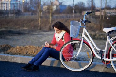 Kız Park edilmiş bir bisikletin yanında oturuyor. Telefonda bir şey izliyorum. Bahar döngüsünde istirahat.
