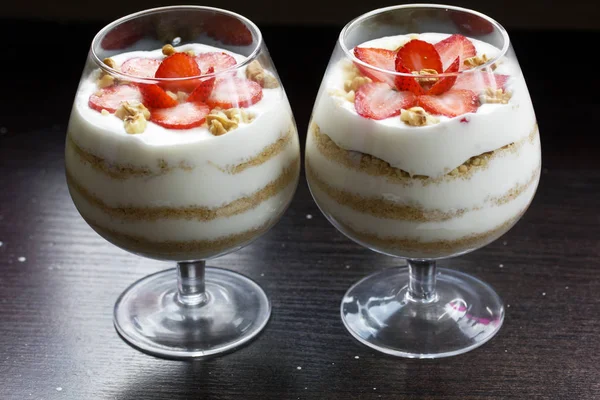 Dessert in a glass goblet. Layers laid biscuit crumbs and cream. Decorated with strawberry slices. With the addition of walnut. On a dark background. — Stock Photo, Image