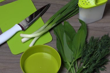 Taze yeşillikler düzgünce masanın üzerine serilir. Kokulu soğan, dereotu ve yabani sarımsak. Salata, tuzlu mahzen, kesme tahtası ve mutfak bıçağı için konteynerin yanında.