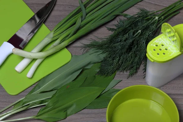 Verduras frescas cuidadosamente colocadas en la mesa. Cebollas aromáticas, eneldo y ajo silvestre. Cerca del contenedor para ensalada, salero, tabla de cortar y cuchillo de cocina . — Foto de Stock