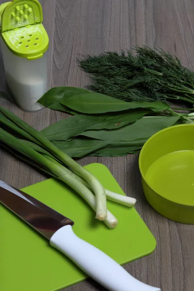 新鲜的绿色整齐地摆放在桌面上。洋葱、萝卜和野生大蒜。靠近沙拉、盐窖、切割板和菜刀的容器. — 图库照片