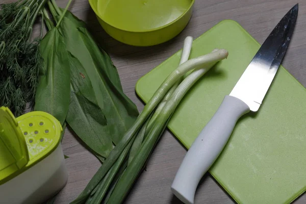 Verduras frescas cuidadosamente colocadas en la mesa. Cebollas aromáticas, eneldo y ajo silvestre. Cerca del contenedor para ensalada, salero, tabla de cortar y cuchillo de cocina . — Foto de Stock