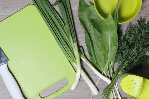 Verduras frescas cuidadosamente colocadas en la mesa. Cebollas aromáticas, eneldo y ajo silvestre. Cerca del contenedor para ensalada, salero, tabla de cortar y cuchillo de cocina . — Foto de Stock