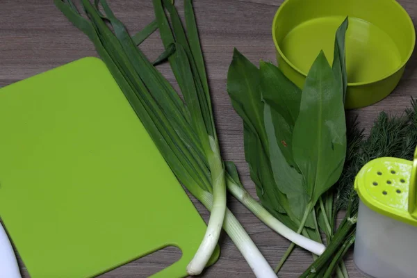 Verduras frescas cuidadosamente colocadas en la mesa. Cebollas aromáticas, eneldo y ajo silvestre. Cerca del contenedor para ensalada, salero, tabla de cortar y cuchillo de cocina . — Foto de Stock