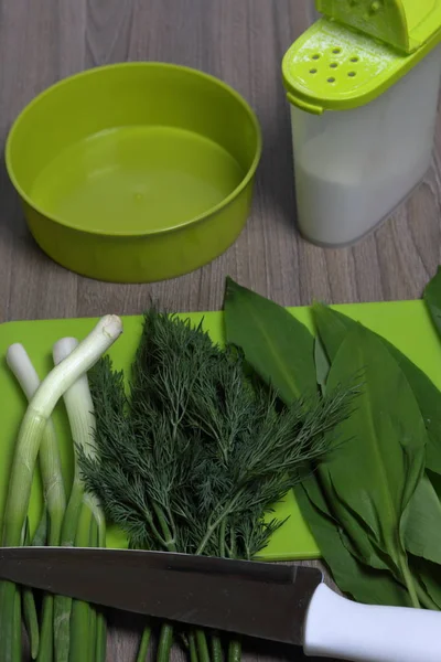 Verduras frescas cuidadosamente colocadas en la mesa. Cebollas aromáticas, eneldo y ajo silvestre. Cerca del contenedor para ensalada, salero, tabla de cortar y cuchillo de cocina . — Foto de Stock