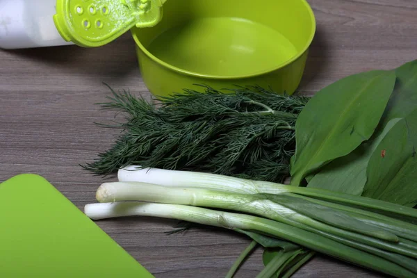 Verduras frescas cuidadosamente colocadas en la mesa. Cebollas aromáticas, eneldo y ajo silvestre. Cerca del contenedor para ensalada, salero y tabla de cortar . — Foto de Stock