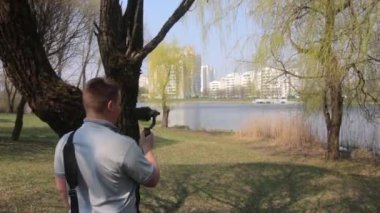 Genç bir adam parkta bir gölün kıyısında duruyor ve bir akıllı telefon üzerinde video çekiyor. Elektronik stabilizatör kullanan pürüzsüz çerçeveler için.