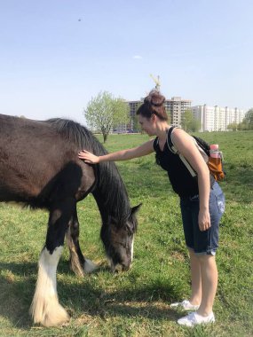 Kız bir atı okşar. Sırt çantasının arkasında, kulaklarda kulaklıklar var..