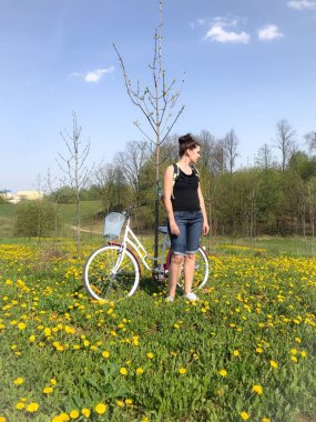 Bir kız bahar çayırında bisikletin yanında duruyor. Sırt çantasının arkasında. Çayırda birçok çiçekli karahindiba var..