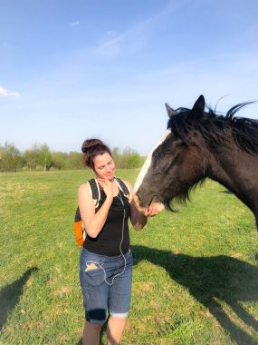 Bahar çayırındaki kız atı besliyor. Sırt çantasının arkasında, kulaklarda kulaklıklar var..