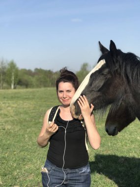 Bahar çayırındaki kız atı besliyor. Sırt çantasının arkasında, kulaklarda kulaklıklar var..