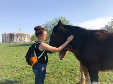 Bahar çayırındaki kız atı besliyor. Sırt çantasının arkasında, kulaklarda kulaklıklar var..