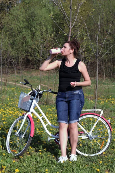 The girl leans on a parked bike. Rest on the spring cycle. The girl drinks water from a bottle.