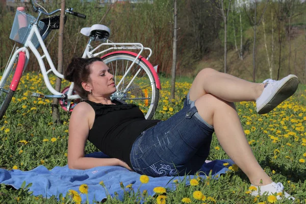 La chica está descansando en el prado de primavera. Se acuesta en la colcha. Muy cerca hay una bicicleta. Los dientes de león están floreciendo, la hierba joven está creciendo . —  Fotos de Stock