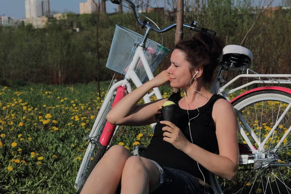 Das Mädchen ruht sich auf der Frühlingswiese aus. Er sitzt neben dem Fahrrad, isst Eis und hört Musik. Löwenzahn blüht, junges Gras wächst. — Stockfoto