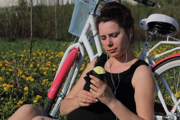 Das Mädchen ruht sich auf der Frühlingswiese aus. Er sitzt neben dem Fahrrad, isst Eis und hört Musik. Löwenzahn blüht, junges Gras wächst. — Stockfoto