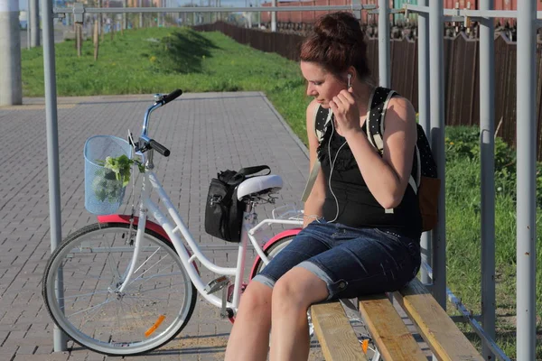 Ein Mädchen sitzt auf einer Bank und hört Musik. In der Nähe liegt ihr Fahrrad. Im Korb liegt ein Strauß Blumen. — Stockfoto