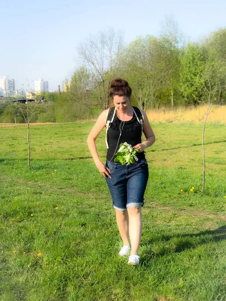 Geht das Mädchen durch eine Frühlingswiese und hält einen Blumenstrauß in den Händen. hinter dem Rucksack, Kopfhörer in den Ohren. — Stockfoto