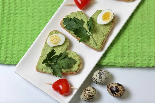 Sandwiches zum Frühstück. herzförmige Brotscheiben mit gemahlener Avocado beschmiert. dekoriert mit gekochten Wachteleiern, Petersilie und Tomaten. entfaltet auf einem weißen rechteckigen Teller. — Stockfoto