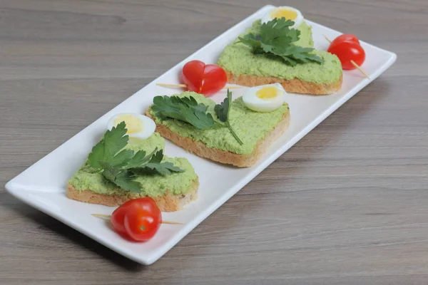 Sandwiches for breakfast. Heart-shaped bread slices smeared with ground avocado. Decorated with boiled quail eggs, parsley and tomatoes. Unfolded on a white rectangular plate.