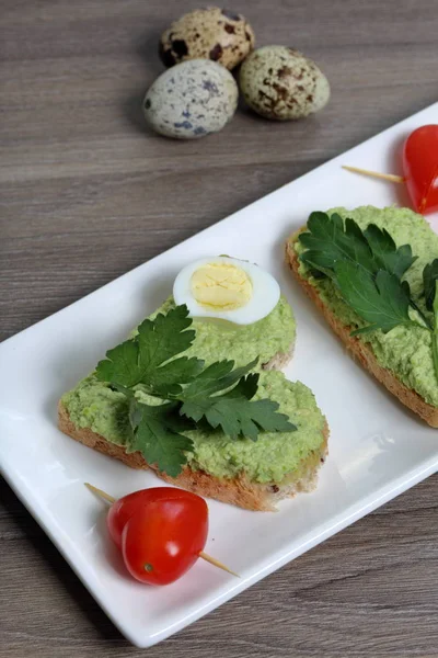 Sandwiches for breakfast. Heart-shaped bread slices smeared with ground avocado. Decorated with boiled quail eggs, parsley and tomatoes. Unfolded on a white rectangular plate.