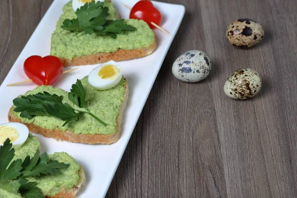 Sandwiches zum Frühstück. herzförmige Brotscheiben mit gemahlener Avocado beschmiert. dekoriert mit gekochten Wachteleiern, Petersilie und Tomaten. entfaltet auf einem weißen rechteckigen Teller. — Stockfoto