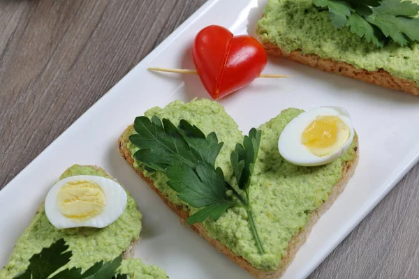 Sandwiches for breakfast. Heart-shaped bread slices smeared with ground avocado. Decorated with boiled quail eggs, parsley and tomatoes. Unfolded on a white rectangular plate.