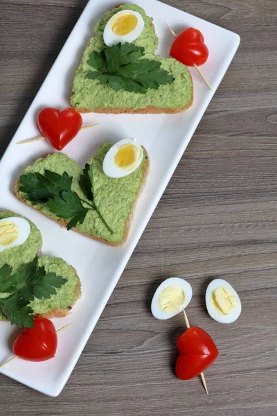 Sandwiches zum Frühstück. herzförmige Brotscheiben mit gemahlener Avocado beschmiert. dekoriert mit gekochten Wachteleiern, Petersilie und Tomaten. entfaltet auf einem weißen rechteckigen Teller. — Stockfoto