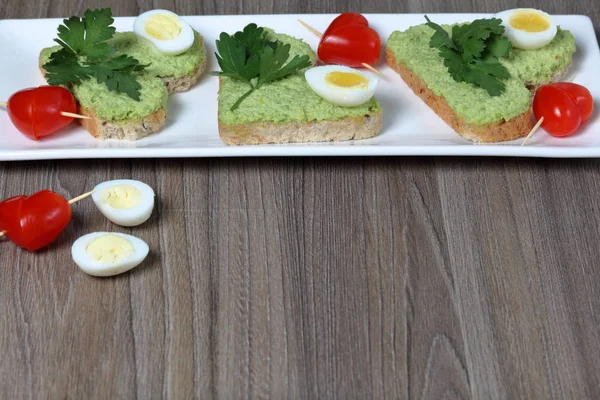 Sandwiches for breakfast. Heart-shaped bread slices smeared with ground avocado. Decorated with boiled quail eggs, parsley and tomatoes. Unfolded on a white rectangular plate.