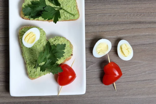 Sandwiches zum Frühstück. herzförmige Brotscheiben mit gemahlener Avocado beschmiert. dekoriert mit gekochten Wachteleiern, Petersilie und Tomaten. entfaltet auf einem weißen rechteckigen Teller. — Stockfoto
