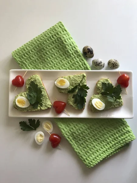 Sandwiches for breakfast. Heart-shaped bread slices smeared with ground avocado. Decorated with boiled quail eggs, parsley and tomatoes. Unfolded on a white rectangular plate. — Stock Photo, Image