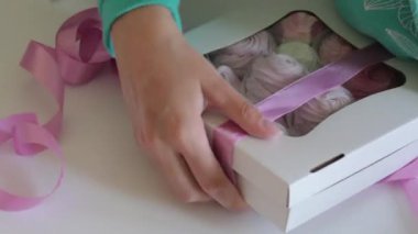 Bir kadın pembe bantla bir kutu şekerleme çalıyor. Kutudaki saydam pencereden görülebilen farklı renklerde marshmallow..