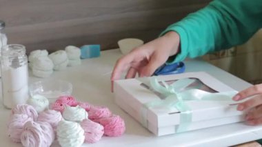 Bir kadın sargılı bir şekerleme kutusu düşünüyor. Örgü yay bağlı. Sıradaki masada paketlenmemiş marşmelovlar var..