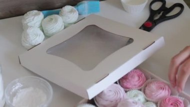 Kadın marşmelovu hediye kutusuna koyuyor. Yakınlarda saydam pencereli bir kutunun kapağı var. Şekerlemeler farklı renklerde, masanın yüzeyine serilmiş...