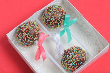 Renkli sprinkles ile çikolata Kek Pops. Kurdele yayı ile dekore edilmiştir. Hediye kutusunda yatıyorlar. Kırmızı arka planda.