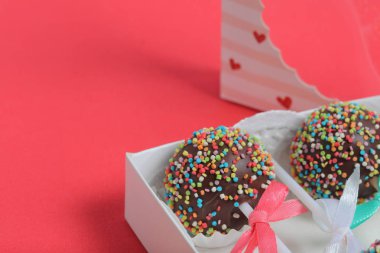 Renkli sprinkles ile çikolata Kek Pops. Kurdele yayı ile dekore edilmiştir. Şeffaf bir pencerenin kapağında bir hediye kutusunda yatıyorlar. Kırmızı arka planda.