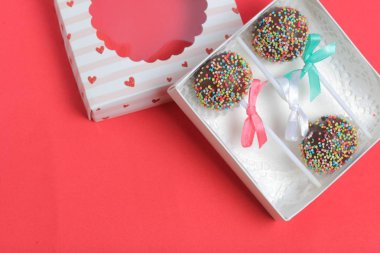 Renkli sprinkles ile çikolata Kek Pops. Kurdele yayı ile dekore edilmiştir. Şeffaf bir pencerenin kapağında bir hediye kutusunda yatıyorlar. Kırmızı arka planda.