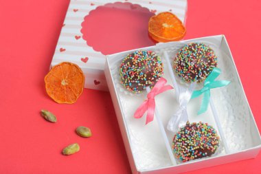 Renkli sprinkles ile çikolata Kek Pops. Kurdele yayı ile dekore edilmiştir. Şeffaf bir pencerenin kapağında bir hediye kutusunda yatıyorlar. Yakında dekorasyon için kurutulmuş meyveler vardır. Kırmızı üzerine 