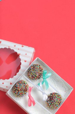 Renkli sprinkles ile çikolata Kek Pops. Kurdele yayı ile dekore edilmiştir. Şeffaf bir pencerenin kapağında bir hediye kutusunda yatıyorlar. Kırmızı arka planda.